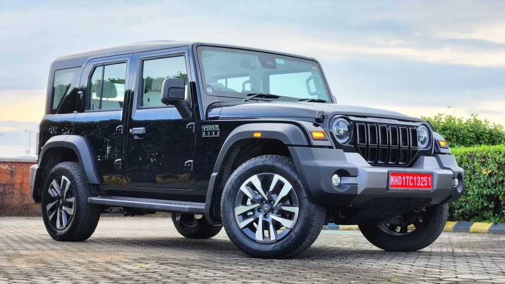 A sleek black 4-door Mahindra Thar, now more appealing with the Mahindra Thar GST Price Cut, is parked on a paved surface with a bright sky in the background, showcasing its extended body and stylish alloy wheels.