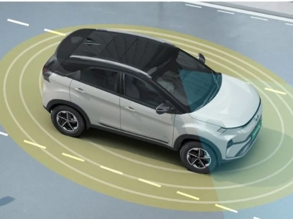 An overhead view of a white Tata Nexon.ev on a road. Yellow and blue animated rings emanate from the car, visually representing its ADAS (Advanced Driver Assistance System) sensors detecting its surroundings.