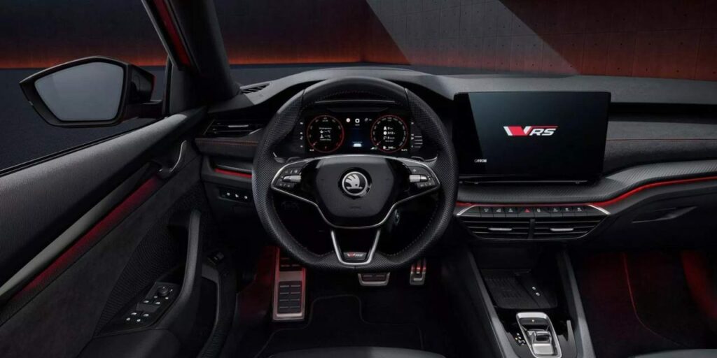 Skoda Octavia RS का हाई-टेक और स्पोर्टी इंटीरियर। इसमें RS लोगो वाला मल्टी-फंक्शनल स्टीयरिंग व्हील, फुली डिजिटल इंस्ट्रूमेंट क्लस्टर, बड़ा टचस्क्रीन इंफोटेनमेंट सिस्टम (जिस पर RS लोगो दिख रहा है), लाल कंट्रास्ट स्टिचिंग और एम्बिएंट लाइटिंग स्पष्ट रूप से देखी जा सकती है। यह कार का ड्राइवर-सेंट्रिक डैशबोर्ड दिखाता है।