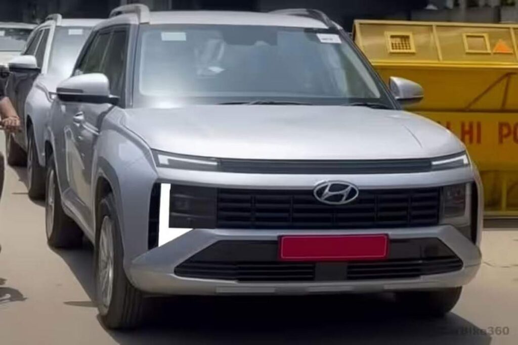 सिल्वर रंग की New-Gen Hyundai Venue (संभवतः बिना ढकी हुई टेस्टिंग यूनिट) का फ्रंट व्यू, जिसमें ऊपर की ओर नई H-आकार की क्षैतिज LED DRLs, बड़ी आयताकार ग्रिल और बम्पर में एकीकृत वर्टिकल हेडलैंप क्लस्टर स्पष्ट रूप से दिखाई दे रहा है। बैकग्राउंड में 'दिल्ली पुलिस' का बैरियर दिख रहा है।