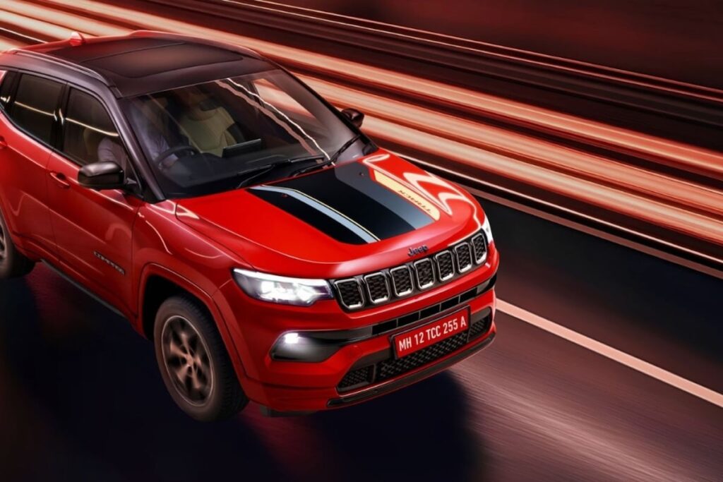 लाल रंग की Jeep Compass Track Edition सड़क पर गतिशील अवस्था में, इसका स्पोर्टी हुड डेकल और आकर्षक डिज़ाइन उजागर कर रही है, जिसकी Jeep Compass Track Edition Price अब ₹26.78 लाख से शुरू होती है।