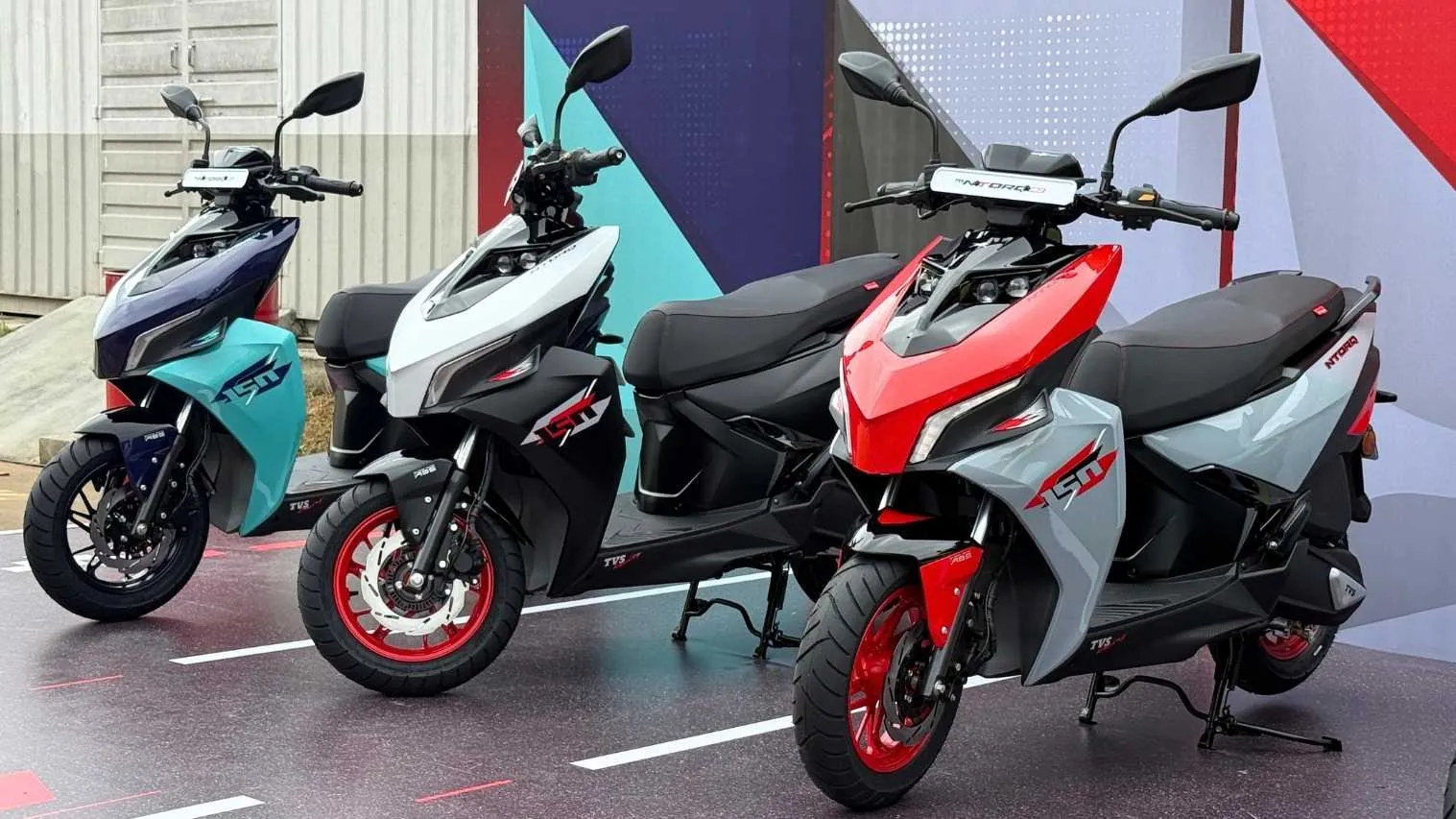तीन अलग-अलग रंग (नीला, सफेद, लाल-ग्रे) की TVS Ntorq 150 स्कूटर लॉन्च इवेंट में खड़ी हैं, जो TVS Q2 FY26 Sales Record में स्कूटर सेगमेंट की 17% वृद्धि को दर्शाती हैं।